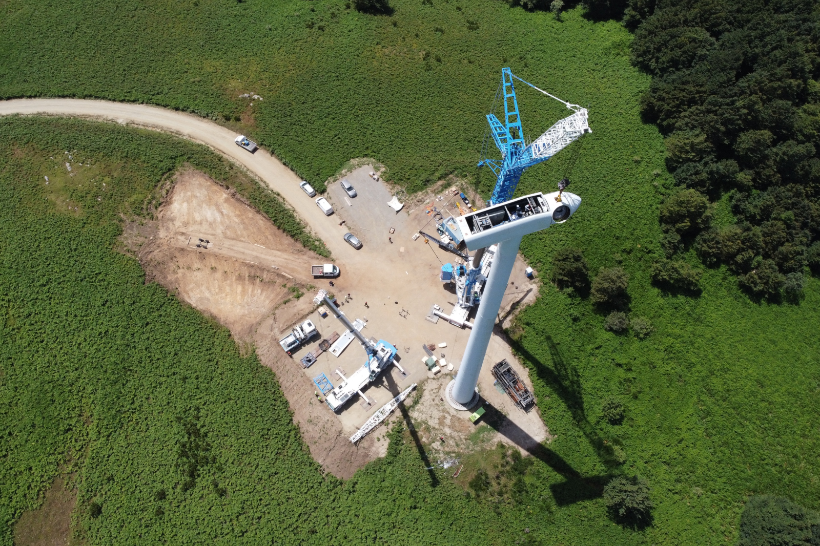 An aerial view of a wind turbine being assembled with a blue crane, parked work vehicles on a cleared site, surrounded by green fields and trees.