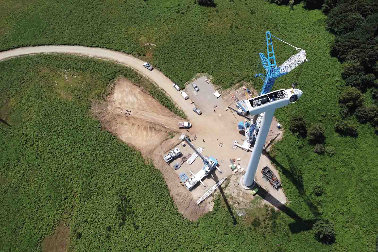 White cylindrical tower with a nacelle stands beside a blue crane on a dirt clearing amid bright green fields, with several white vehicles.
