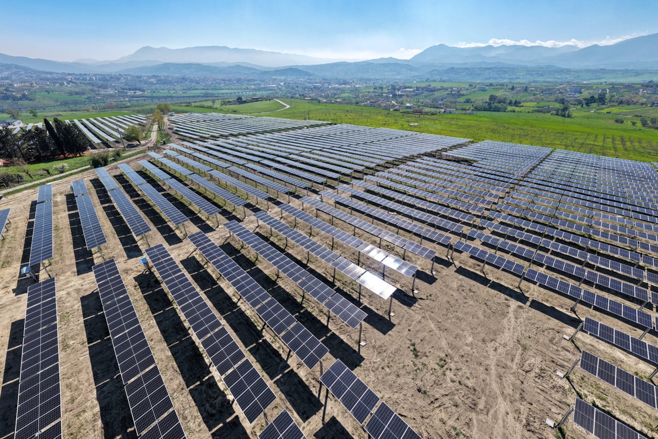 Vast rows of dark blue solar panels on metal frames stretch across brown soil toward green fields and distant mountains beneath a clear blue sky.