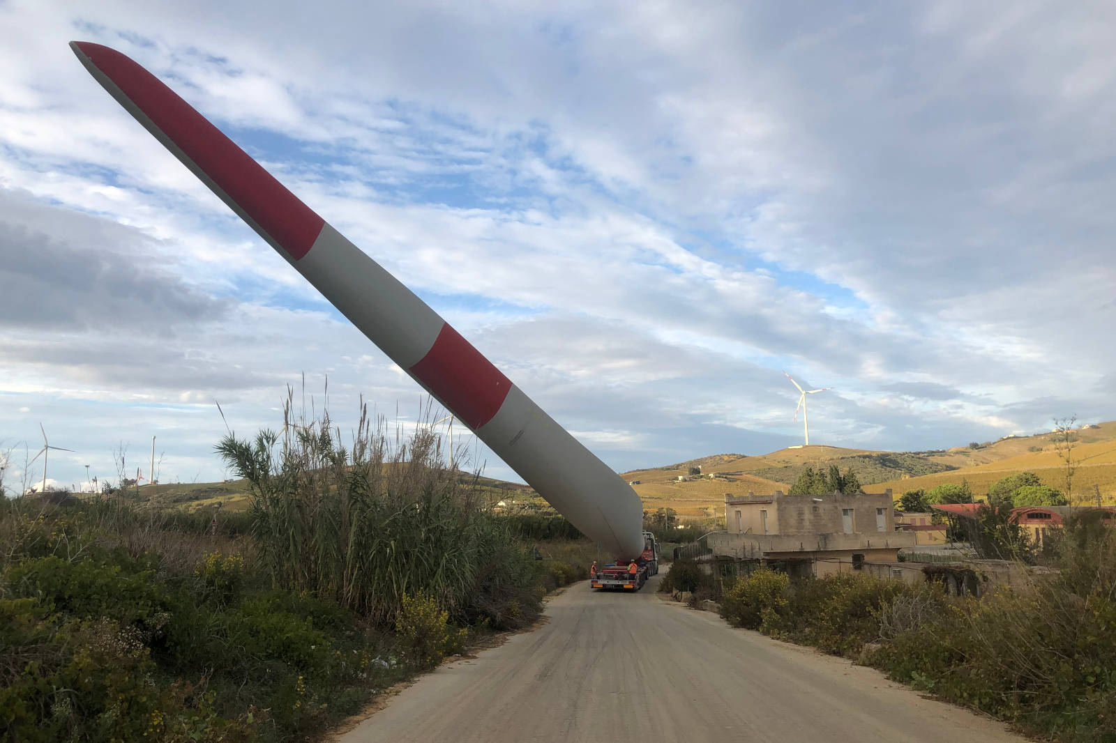 A truck transports a large wind turbine blade down a road. Hills and additional wind turbines are visible in the background.