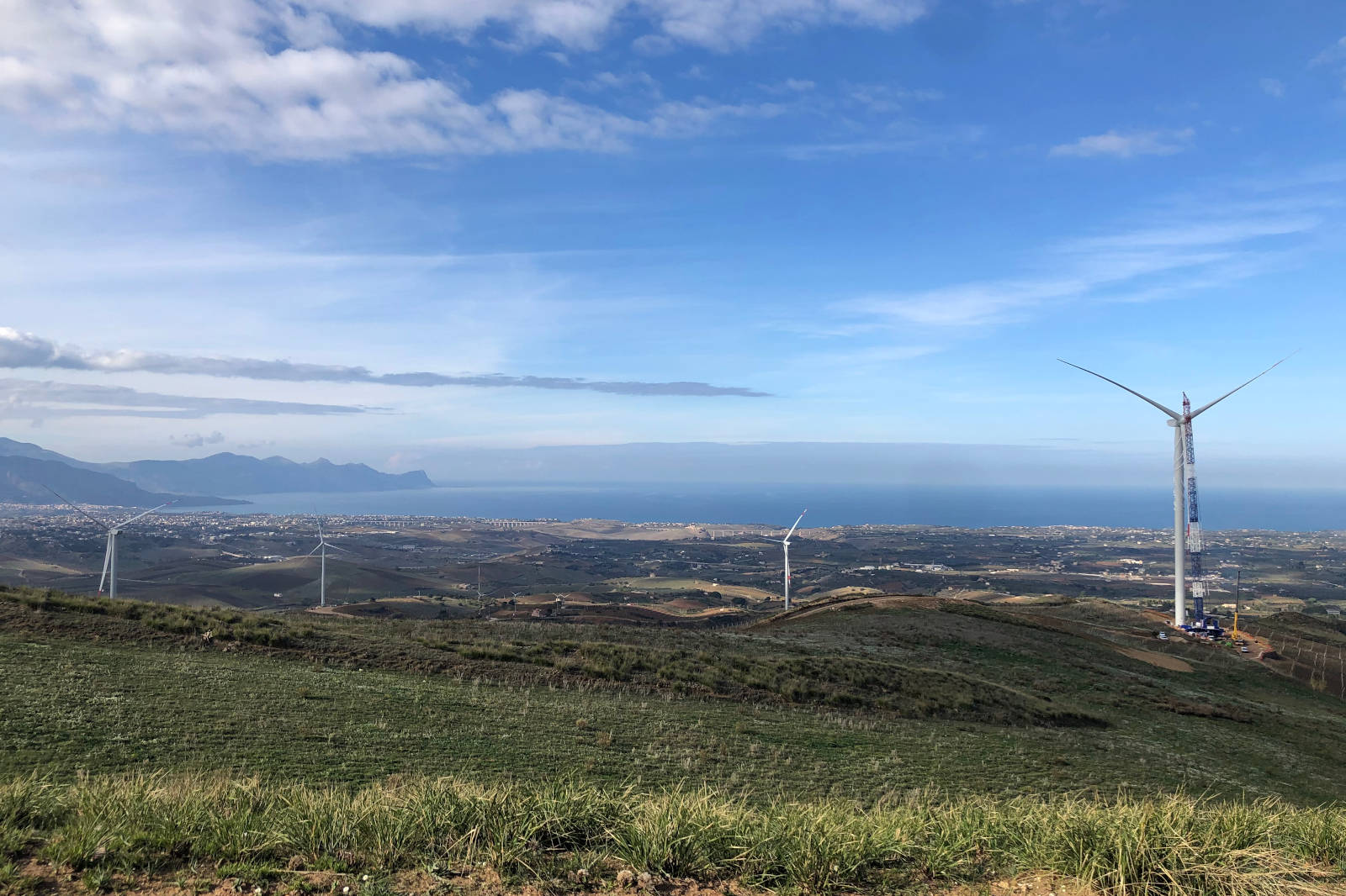 A landscape featuring wind turbines, hills, and the sea in the background under a blue sky.