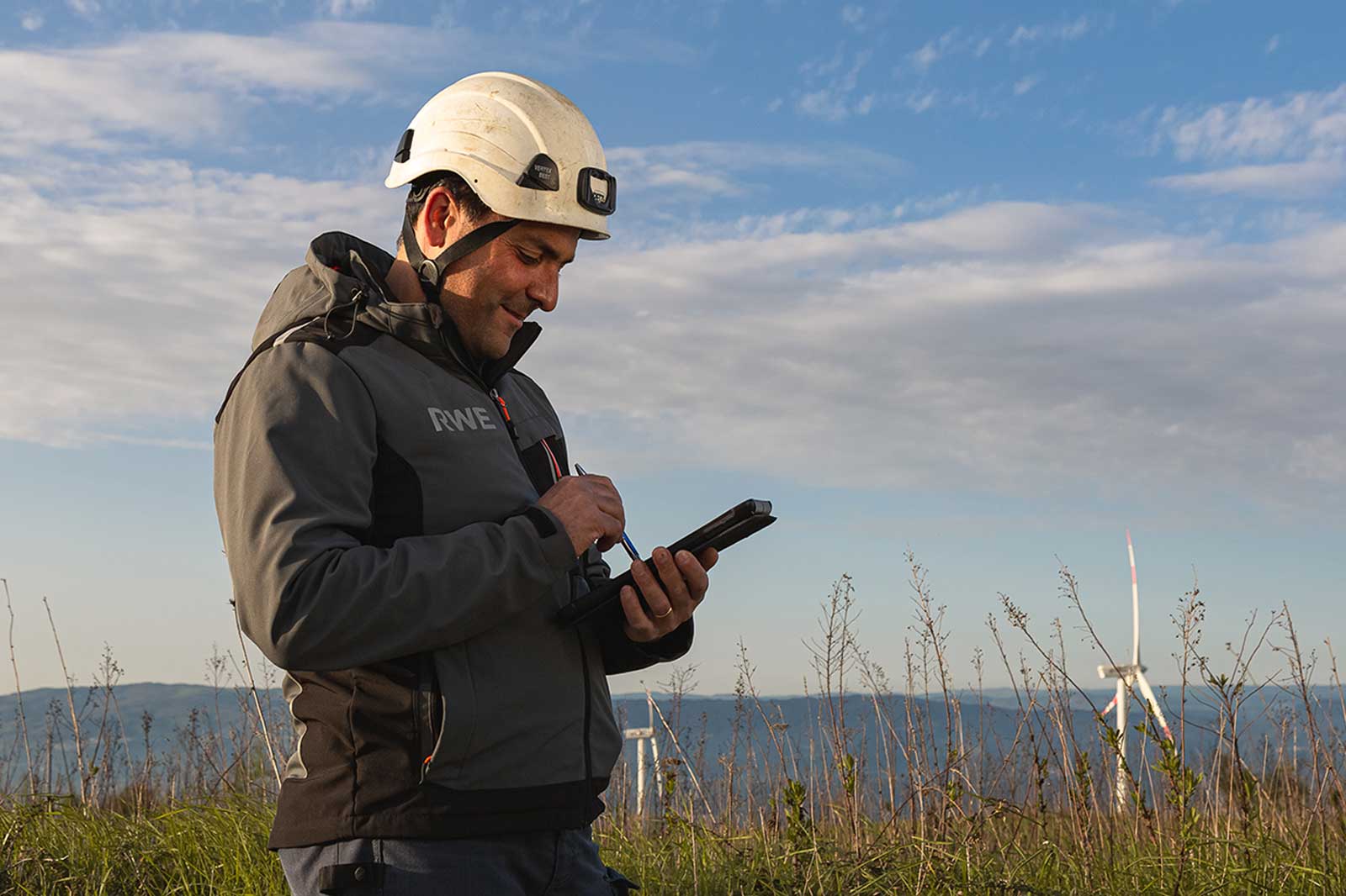 Un lavoratore con un casco utilizza un tablet all'aperto vicino a turbine eoliche sotto un cielo blu con nuvole sparse.
