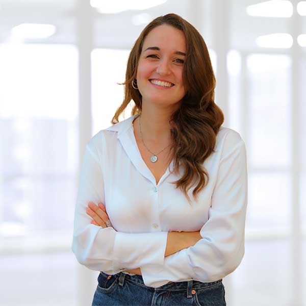 Una donna con capelli lunghi e mossi sta in piedi con le braccia incrociate, indossando una camicia bianca e jeans in un ufficio luminoso.