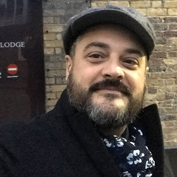 Un uomo con un cappotto scuro e un berretto è in piedi contro un muro di mattoni, indossando una sciarpa a fantasia.