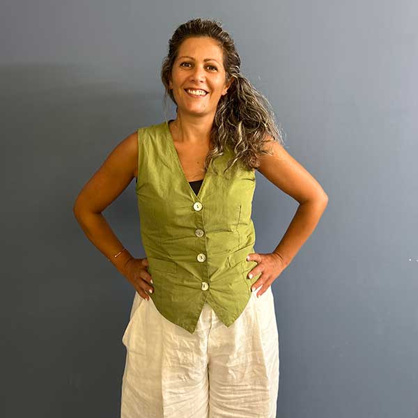 Una donna sta in piedi con sicurezza indossando un top verde senza maniche con bottoni e pantaloni bianchi su uno sfondo grigio.