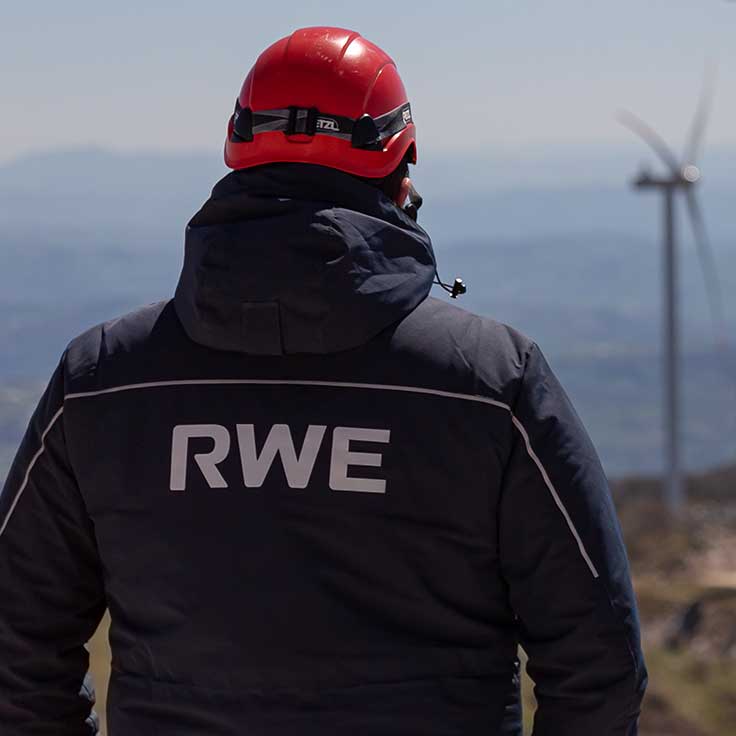 Un lavoratore con un casco rosso e una giacca scura con il logo RWE guarda le turbine eoliche in un paesaggio montano.